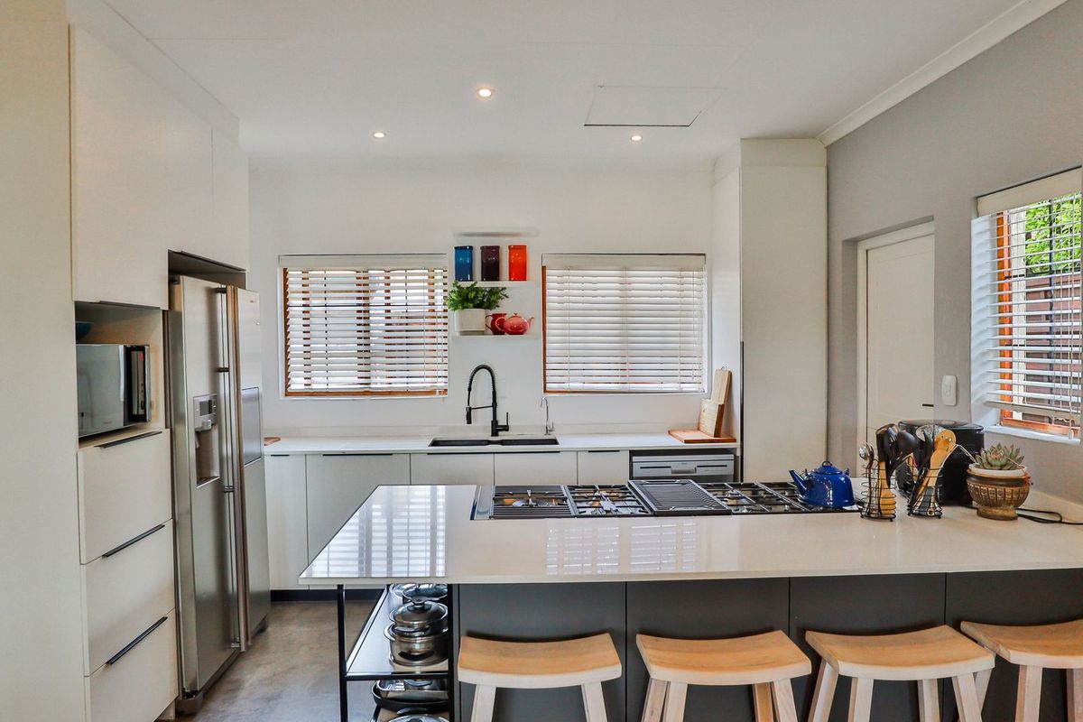 Modern kitchen featuring sleek white cabinetry, stone countertops, a central island with a gas cooktop, and ample natural light through venetian blinds.