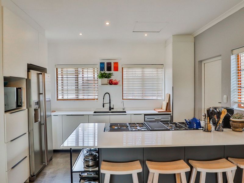 Modern kitchen featuring sleek white cabinetry, stone countertops, a central island with a gas cooktop, and ample natural light through venetian blinds.