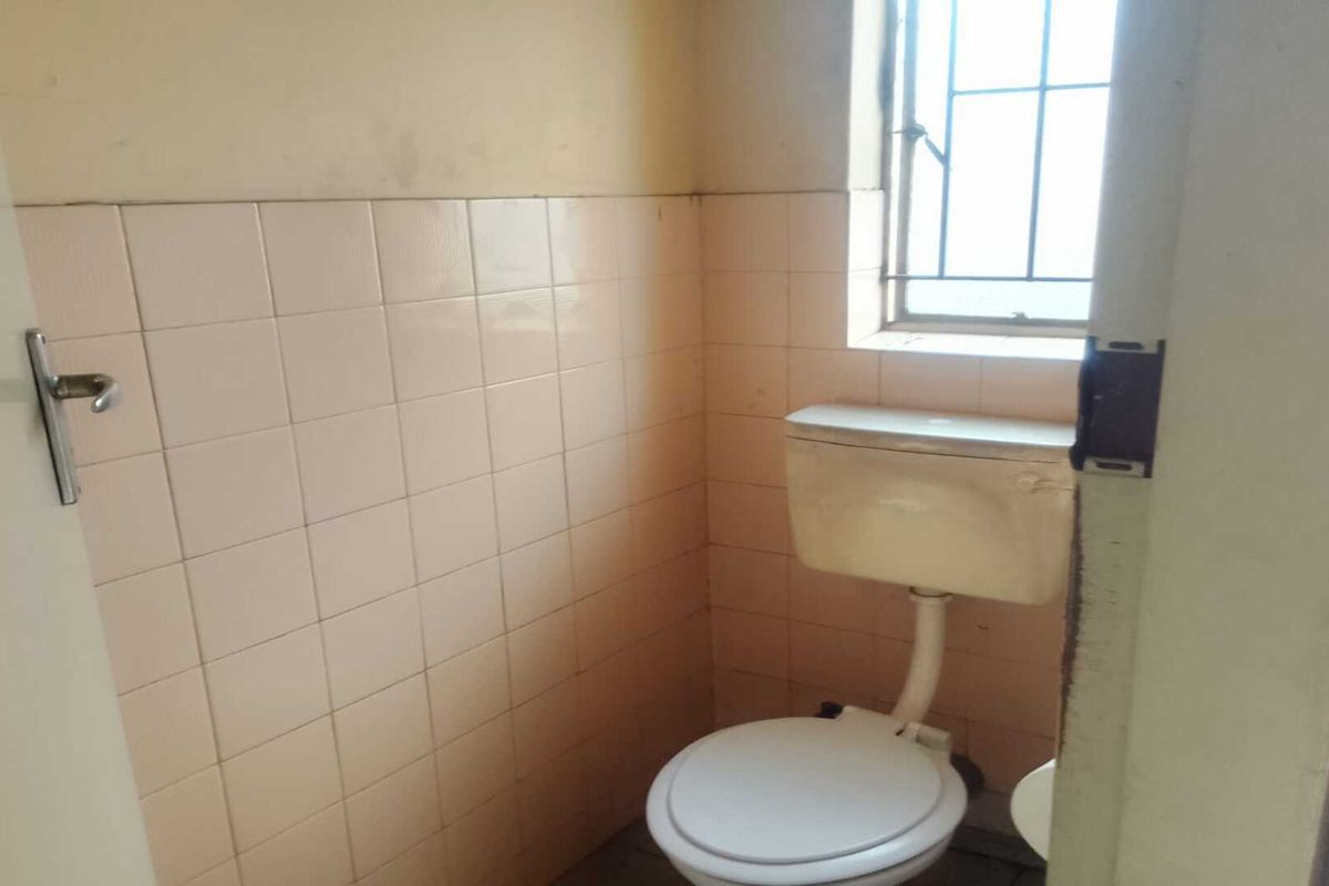 A compact industrial restroom featuring dated wall tiling, a standard toilet suite, and a security-barred window.