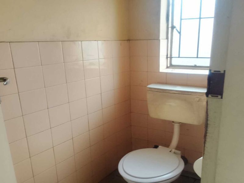 A compact industrial restroom featuring dated wall tiling, a standard toilet suite, and a security-barred window.
