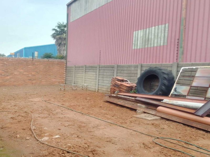 An exterior industrial yard area featuring a corrugated metal warehouse wall, brick perimeter fencing, and an unpaved ground surface.
