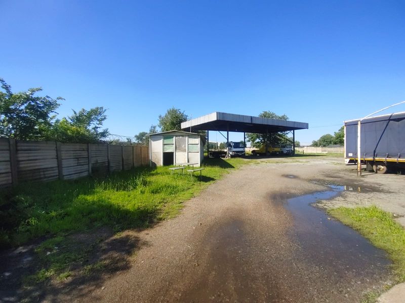 Industrial yard featuring a large covered parking structure, a small utility shed, and perimeter concrete walling on a gravel surface.