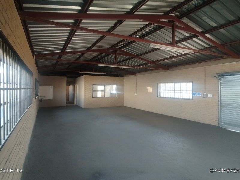 Spacious industrial utility area featuring exposed steel roof trusses, painted concrete flooring, and a secure roller shutter door, suitable for workshop or storage use.