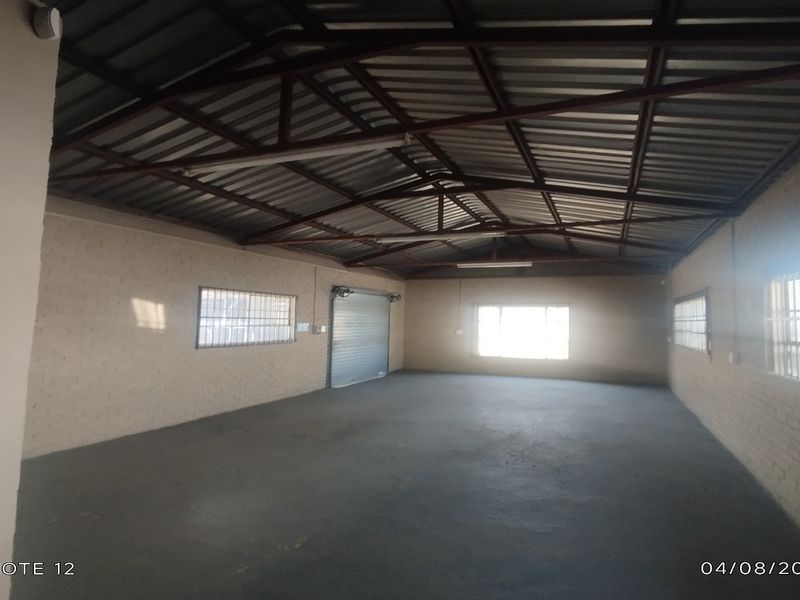 Spacious industrial utility area featuring exposed steel roof trusses, concrete flooring, and secure roller shutter access, suitable for storage or workshop use.
