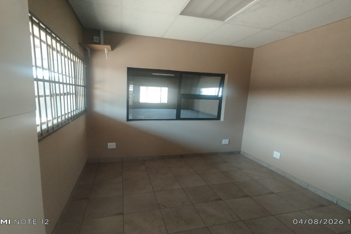 A vacant office space featuring tiled flooring, a drop ceiling, security bars on the window, and an internal window looking into an adjacent area.