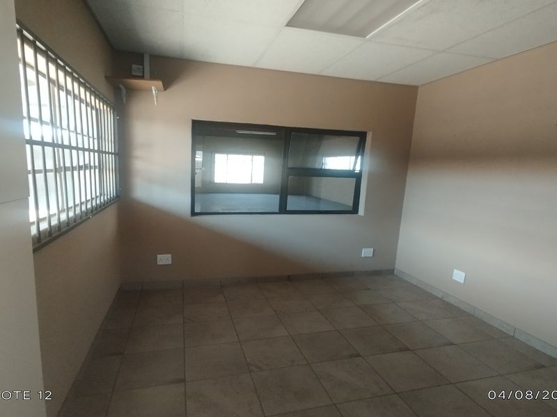 A vacant office space featuring tiled flooring, a drop ceiling, security bars on the window, and an internal window looking into an adjacent area.