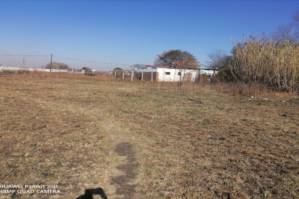 A wide-angle view of a large, undeveloped plot of land featuring dry grass, perimeter fencing, and a small, weathered outbuilding in the distance.
