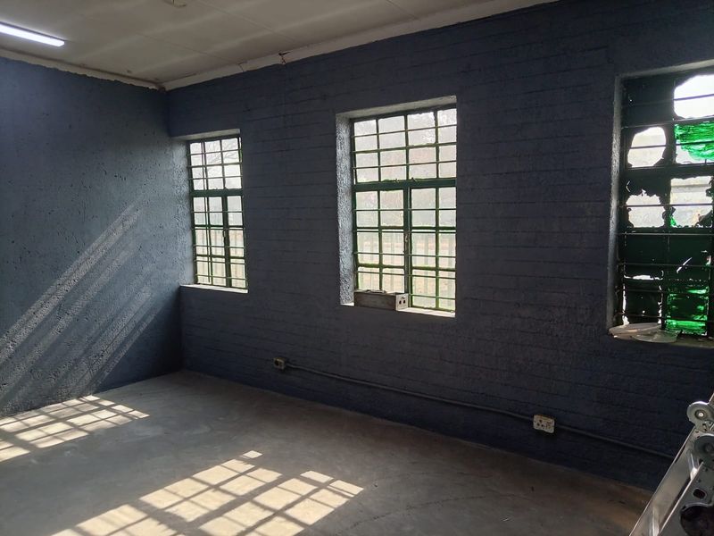 An empty industrial utility space featuring exposed brick walls, surface-mounted electrical conduits, and steel-framed windows with security bars.
