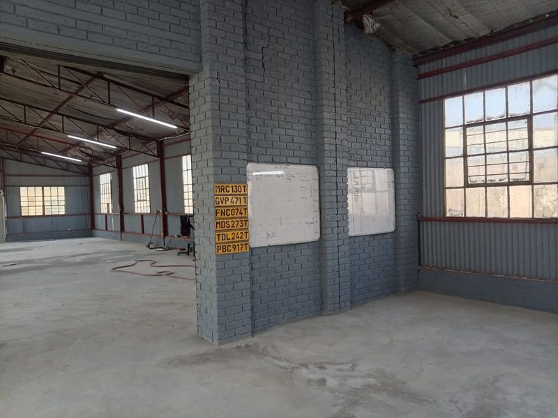 Spacious industrial warehouse interior featuring exposed steel roof trusses, painted brick columns, and large steel-framed windows with concrete flooring.