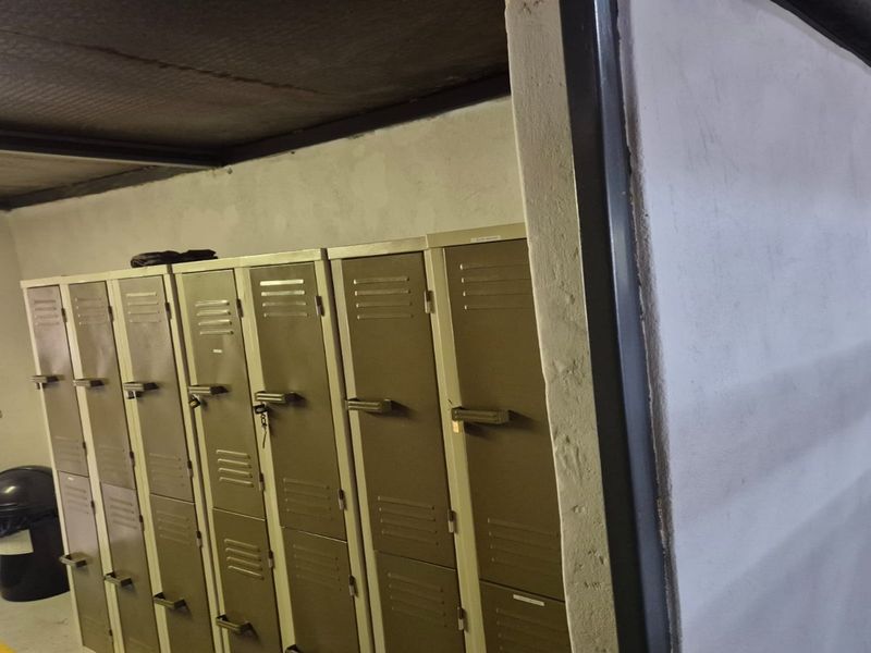 A utility or staff locker room featuring built-in metal storage lockers, exposed steel ceiling beams, and unfinished painted masonry walls.