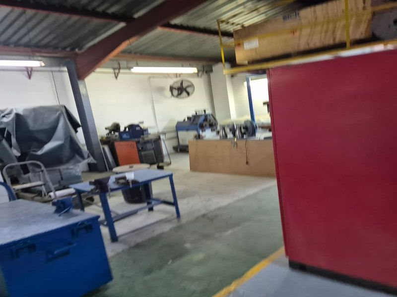 Industrial utility space featuring exposed steel roof structure, concrete flooring, and wall-mounted industrial ventilation fan, suitable for workshop or storage operations.