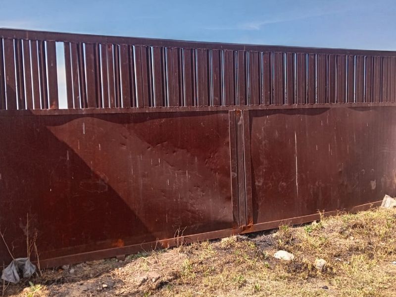 A tall, industrial-style metal perimeter fence and gate showing significant signs of surface rust and structural wear.