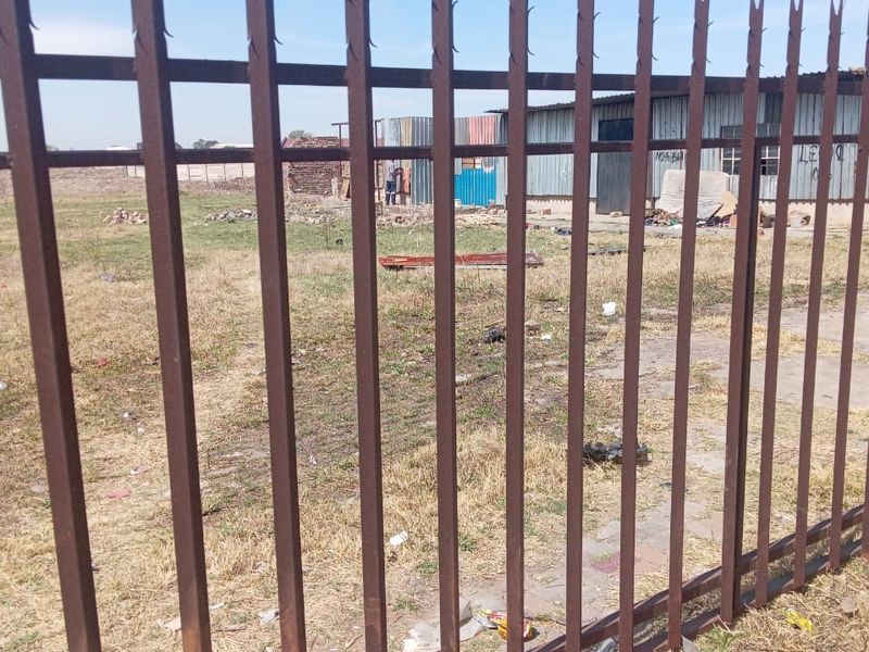 View of an industrial property featuring metal spike perimeter fencing and a corrugated metal structure on a large, unmaintained plot of land.