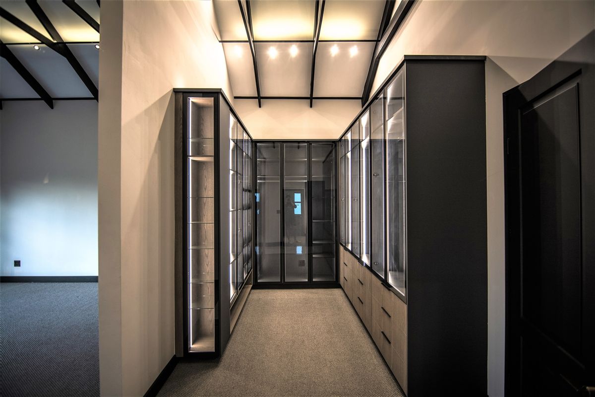 A modern walk-in dressing room featuring custom built-in cabinetry with glass-fronted doors, integrated LED lighting, and a high vaulted ceiling with exposed beams.