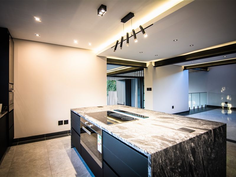 Modern kitchen featuring a large stone island with integrated cooktop and oven, sleek black cabinetry, and contemporary recessed and pendant lighting.