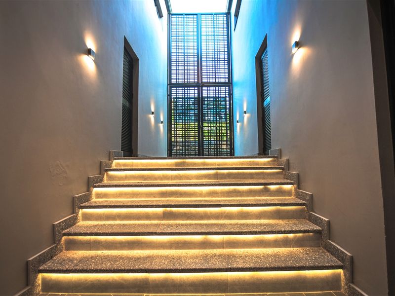 A modern, well-lit entrance featuring a stone staircase with integrated LED lighting, leading up to a large metal security gate and overhead skylight.
