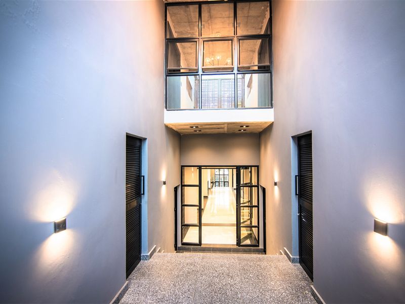 A modern, high-ceilinged hallway featuring terrazzo flooring, black metal-framed glass doors, and sophisticated architectural lighting, creating a clean and contemporary transition space.