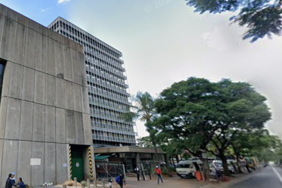 A multi-story office building featuring a concrete facade and glass curtain walling, situated along a street with mature trees.