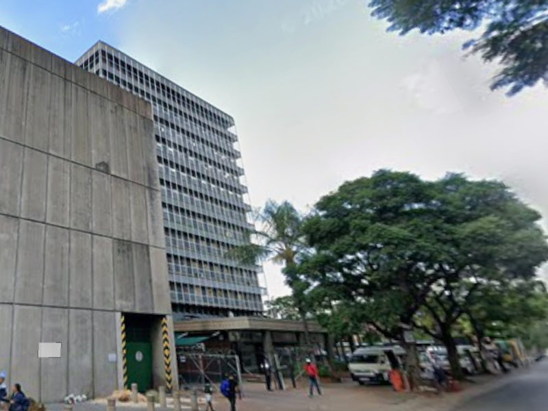 A multi-story office building featuring a concrete facade and glass curtain walling, situated along a street with mature trees.