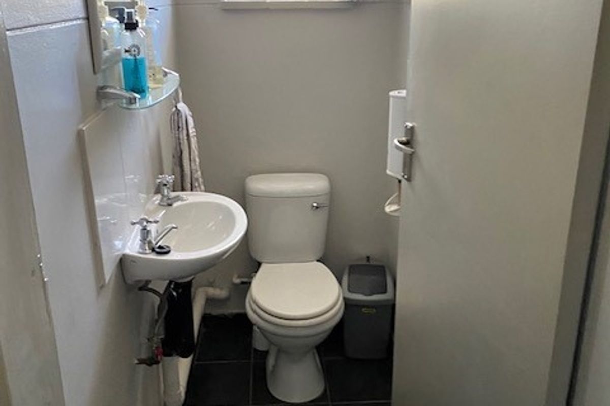 A compact office restroom featuring a wall-mounted basin, a standard toilet, and dark floor tiling with a security-barred window.