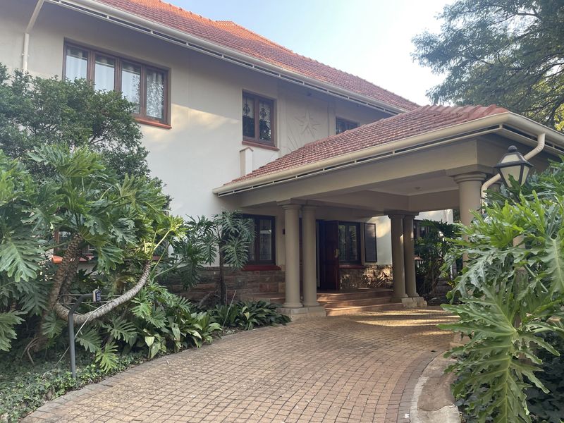 Front elevation of a classic residence featuring a covered entrance portico, stone detailing, and a paved driveway surrounded by established garden foliage.