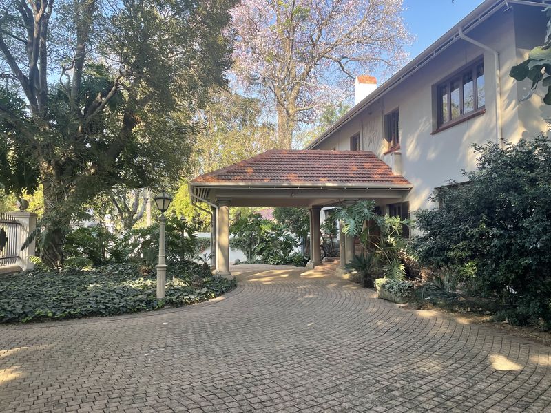 A well-maintained front entrance featuring a brick-paved driveway leading to a covered porte-cochere with stone columns, surrounded by mature trees and landscaped garden beds.