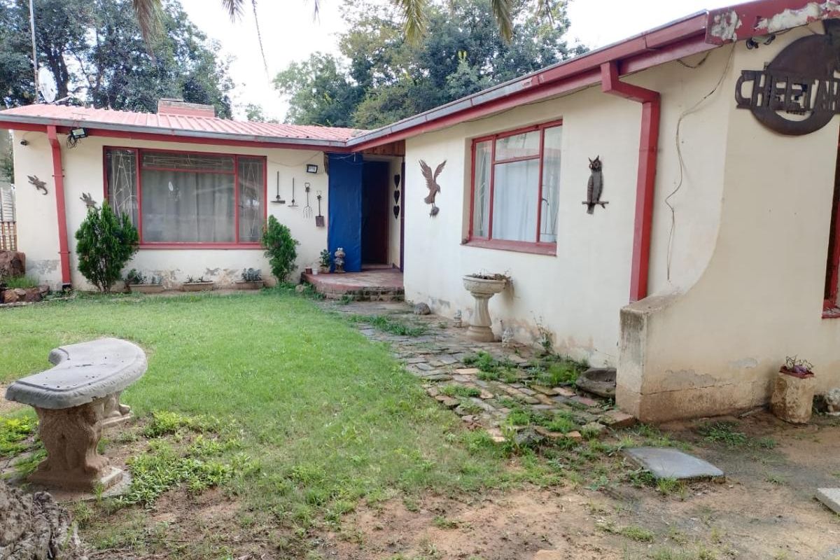 Exterior view of a single-story house featuring a red metal roof, stone pathway, and garden area requiring maintenance and cosmetic updates.