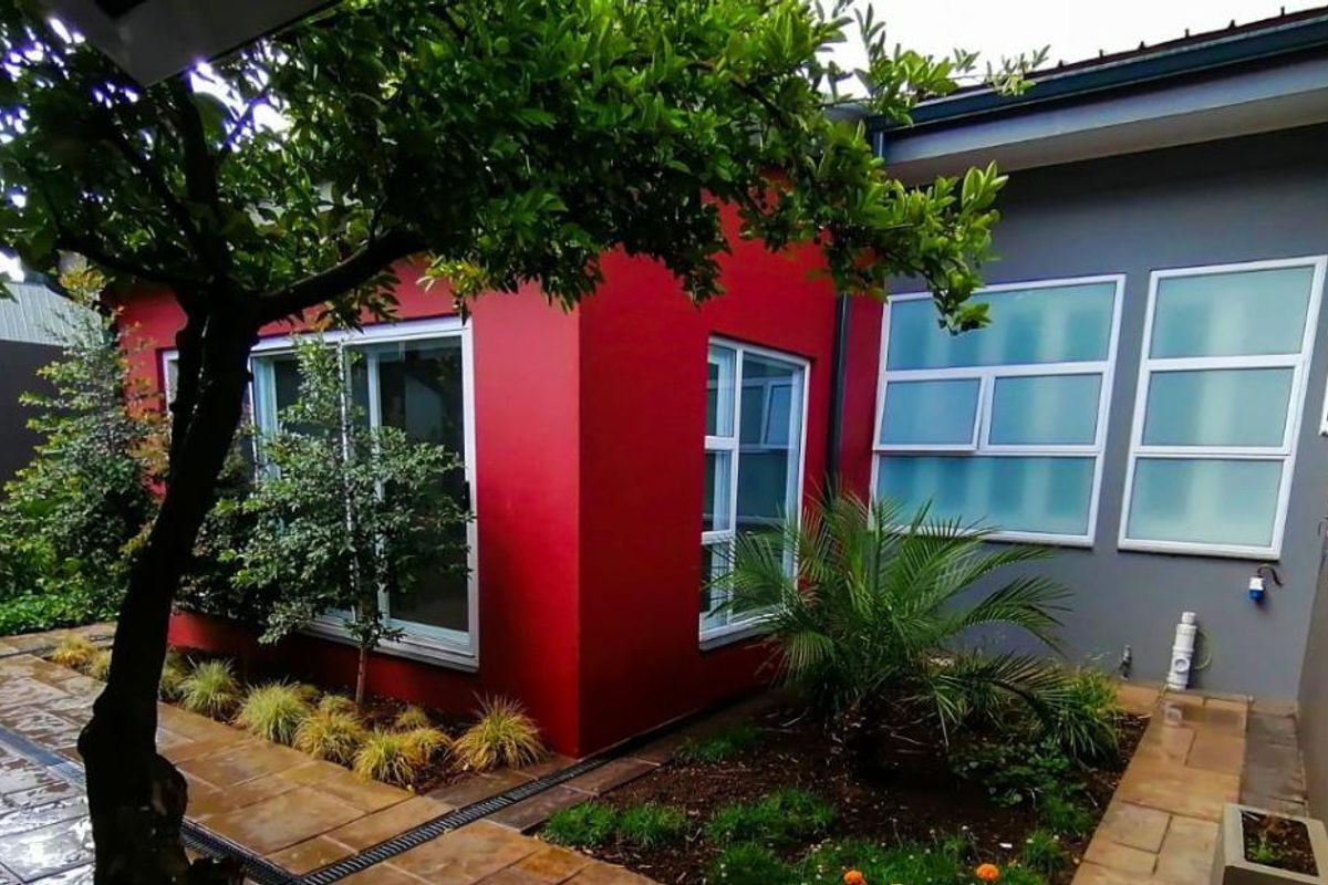 A well-maintained rear exterior view featuring a vibrant red accent wall, modern aluminium-framed windows, paved courtyard area, and established garden landscaping.
