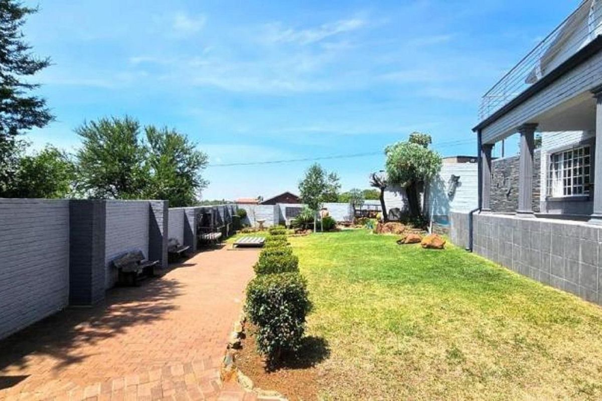 Spacious rear garden featuring a paved walkway, manicured lawn, and a covered patio area with structural columns.
