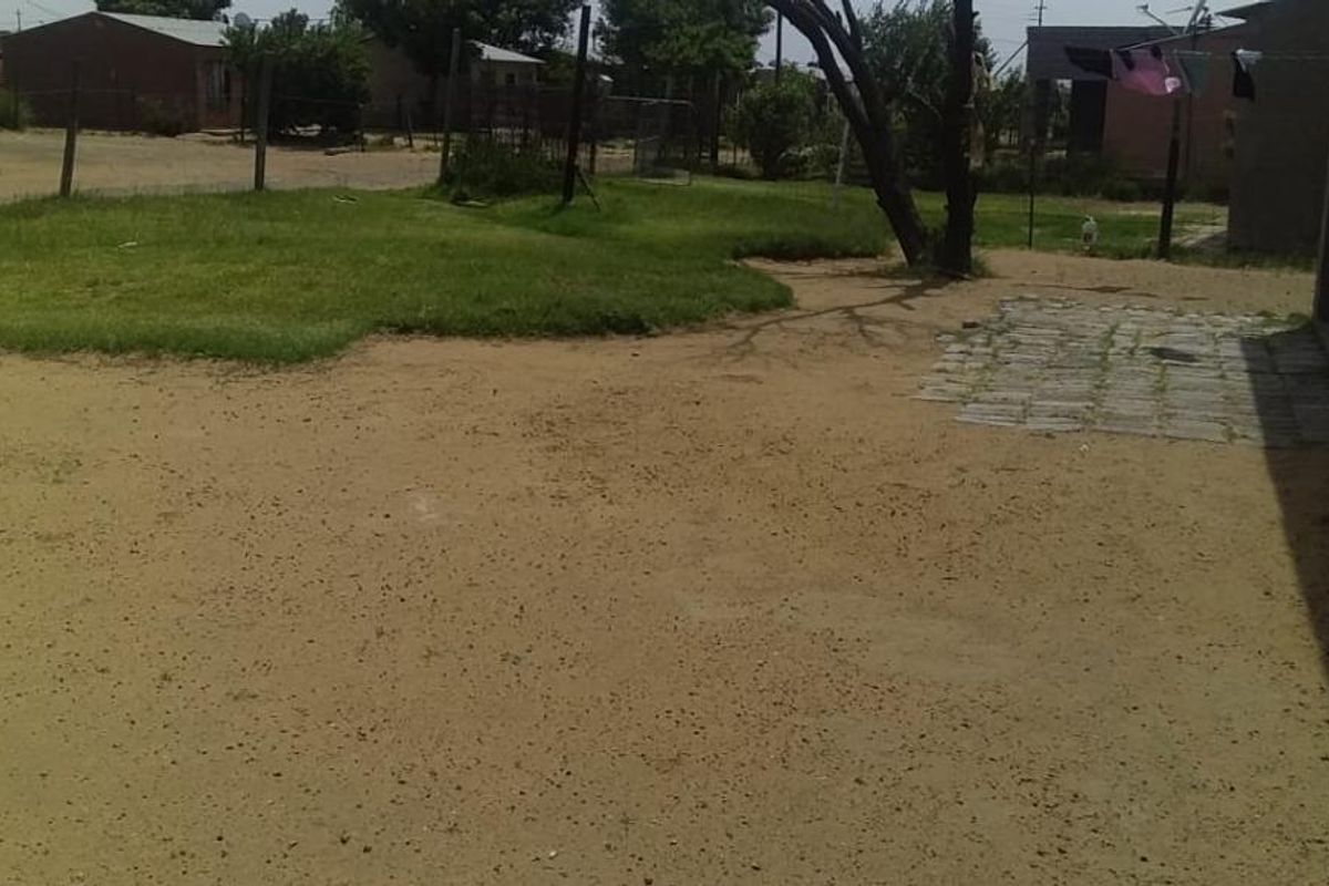 A view of the rear yard area featuring a mix of sandy ground, a small patch of lawn, and a paved section adjacent to the house.