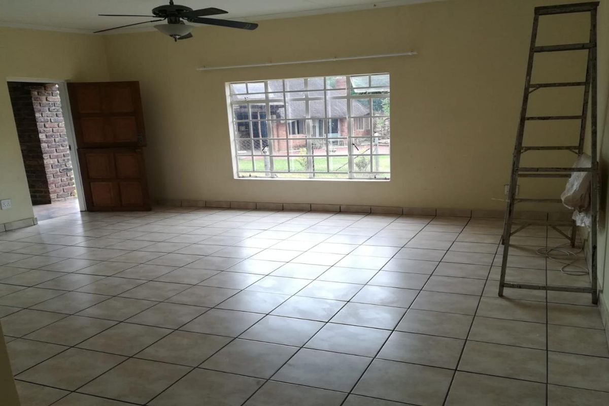 A spacious, empty living room featuring neutral ceramic floor tiles, a ceiling fan, and large windows providing natural light and garden views.