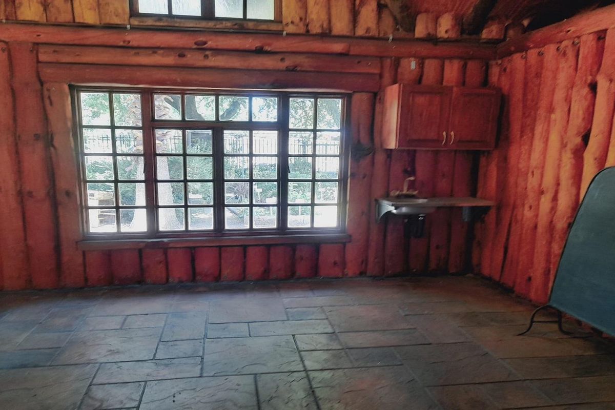 A rustic kitchen area featuring log walls, stone tile flooring, a large lattice window, and a wall-mounted cabinet with a stainless steel sink.