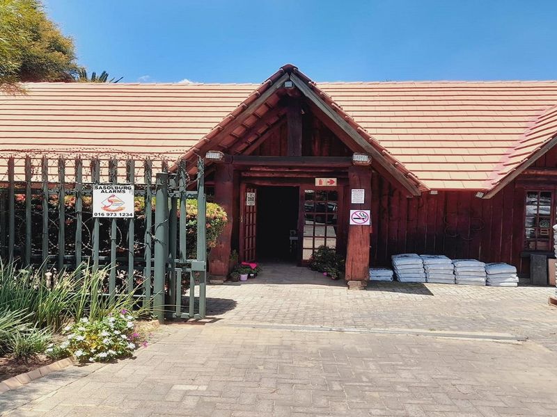 Front exterior view of a timber-clad building with a tiled roof, featuring a paved entrance area and secure perimeter fencing.