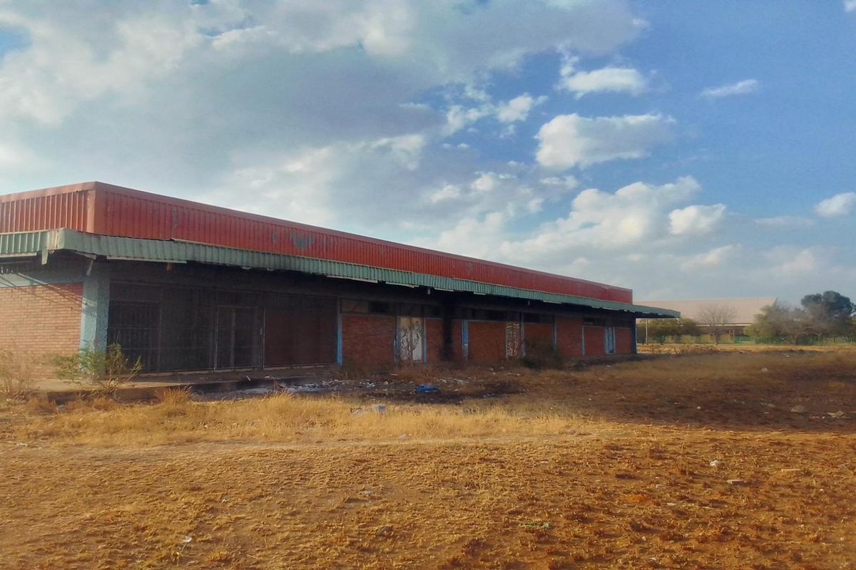 A view of a vacant commercial or industrial structure featuring a brick facade and metal roofing, situated on a large, undeveloped plot of land.