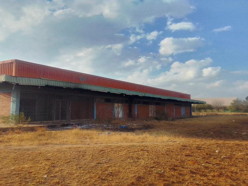 A view of a vacant commercial or industrial structure featuring a brick facade and metal roofing, situated on a large, undeveloped plot of land.
