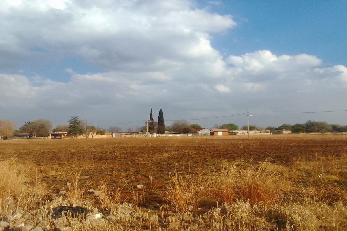 A wide, open residential vacant land plot featuring dry grass and clear views of the surrounding neighborhood and horizon.