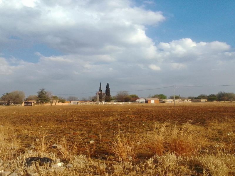 A wide, open residential vacant land plot featuring dry grass and clear views of the surrounding neighborhood and horizon.