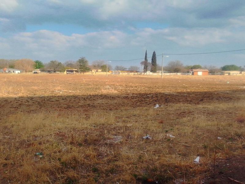 A wide, vacant residential plot of land featuring dry grass and visible utility infrastructure, offering a clear view of the surrounding neighborhood.