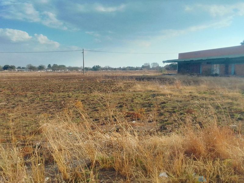 A wide view of a vacant residential plot featuring dry grass, an adjacent brick building, and an overhead utility line.
