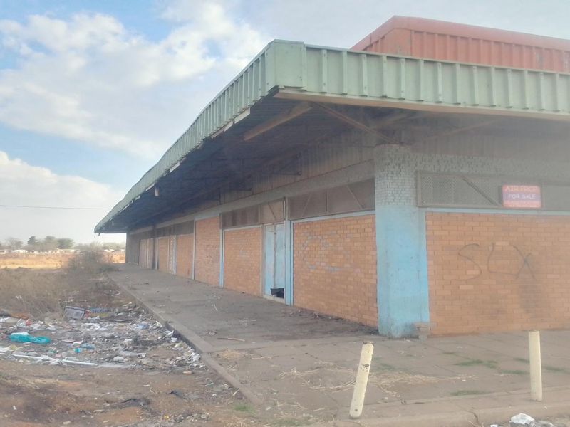 A view of a commercial building exterior featuring a brick facade, metal roof canopy, and concrete walkway, currently showing signs of neglect and debris.
