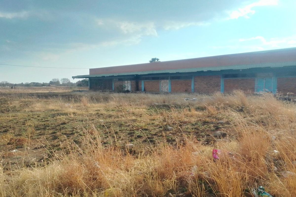 A view of a vacant residential plot featuring a dilapidated brick structure with a metal roof, surrounded by overgrown dry grass.