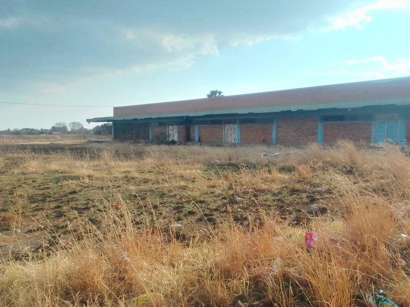A view of a vacant residential plot featuring a dilapidated brick structure with a metal roof, surrounded by overgrown dry grass.