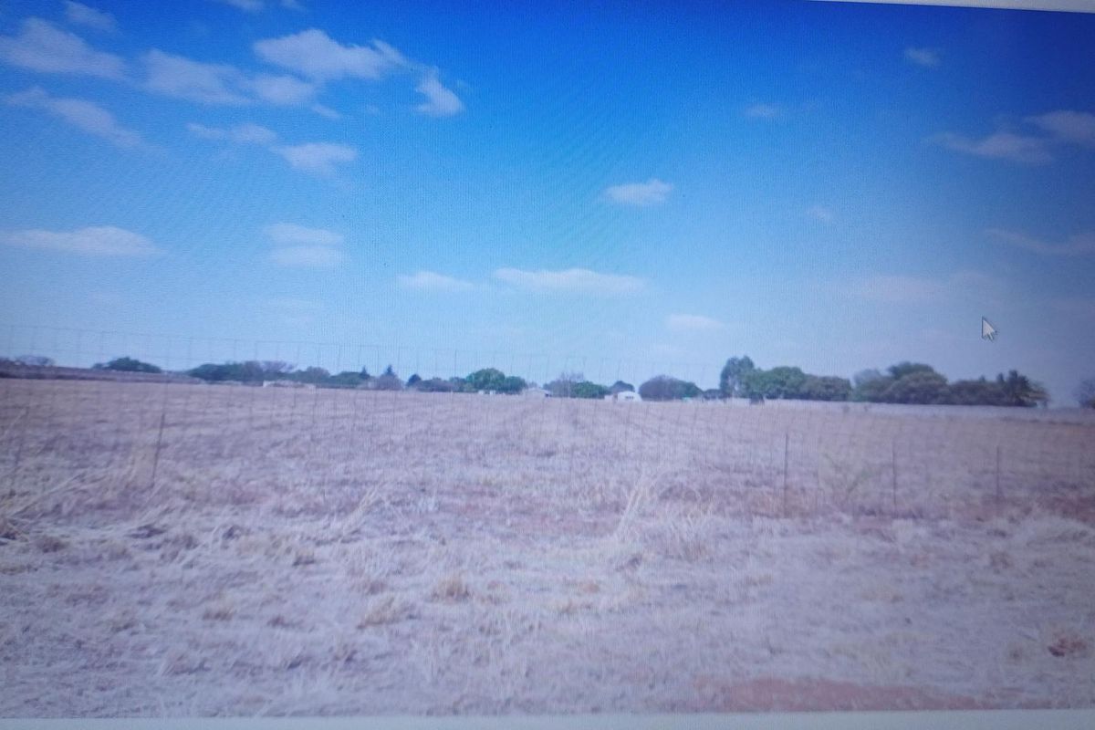 A wide-angle view of a vacant residential plot featuring dry grass and perimeter wire fencing under a clear blue sky.
