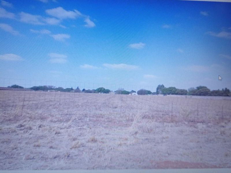 A wide-angle view of a vacant residential plot featuring dry grass and perimeter wire fencing under a clear blue sky.