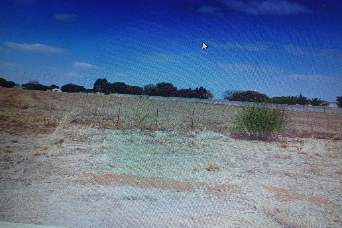 A view of a vacant residential plot featuring dry grass and perimeter wire fencing under a clear blue sky.