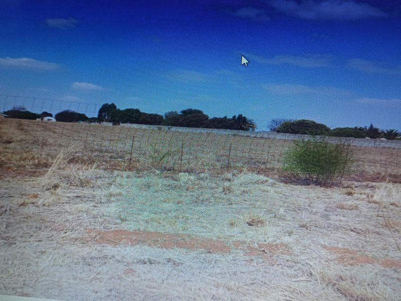 A view of a vacant residential plot featuring dry grass and perimeter wire fencing under a clear blue sky.
