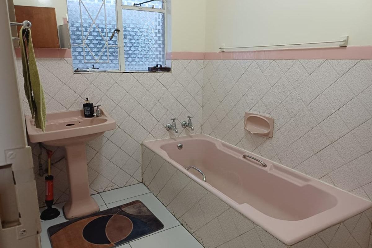 A dated bathroom featuring a pink pedestal basin and matching bathtub, complemented by diamond-patterned wall tiles and a security-grilled window.