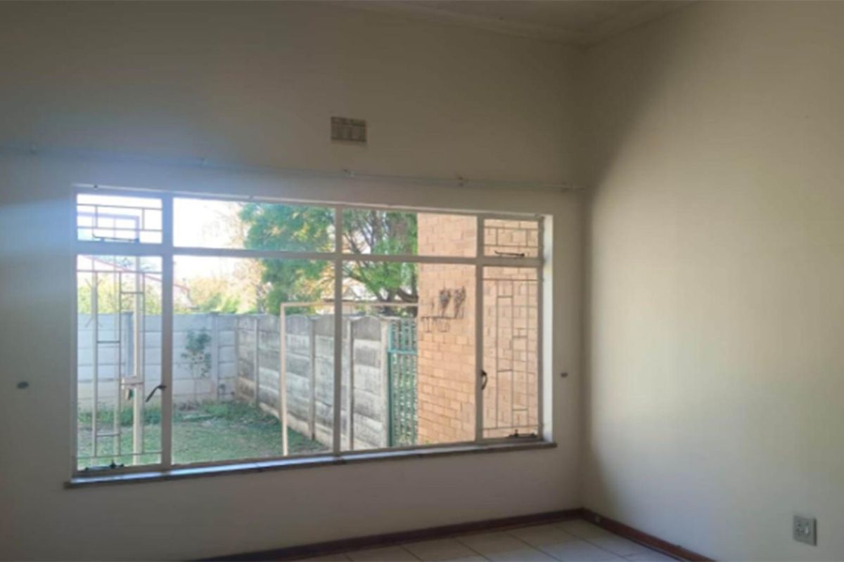 An empty living room featuring large windows with security bars, tiled flooring, and a view of the rear garden area.