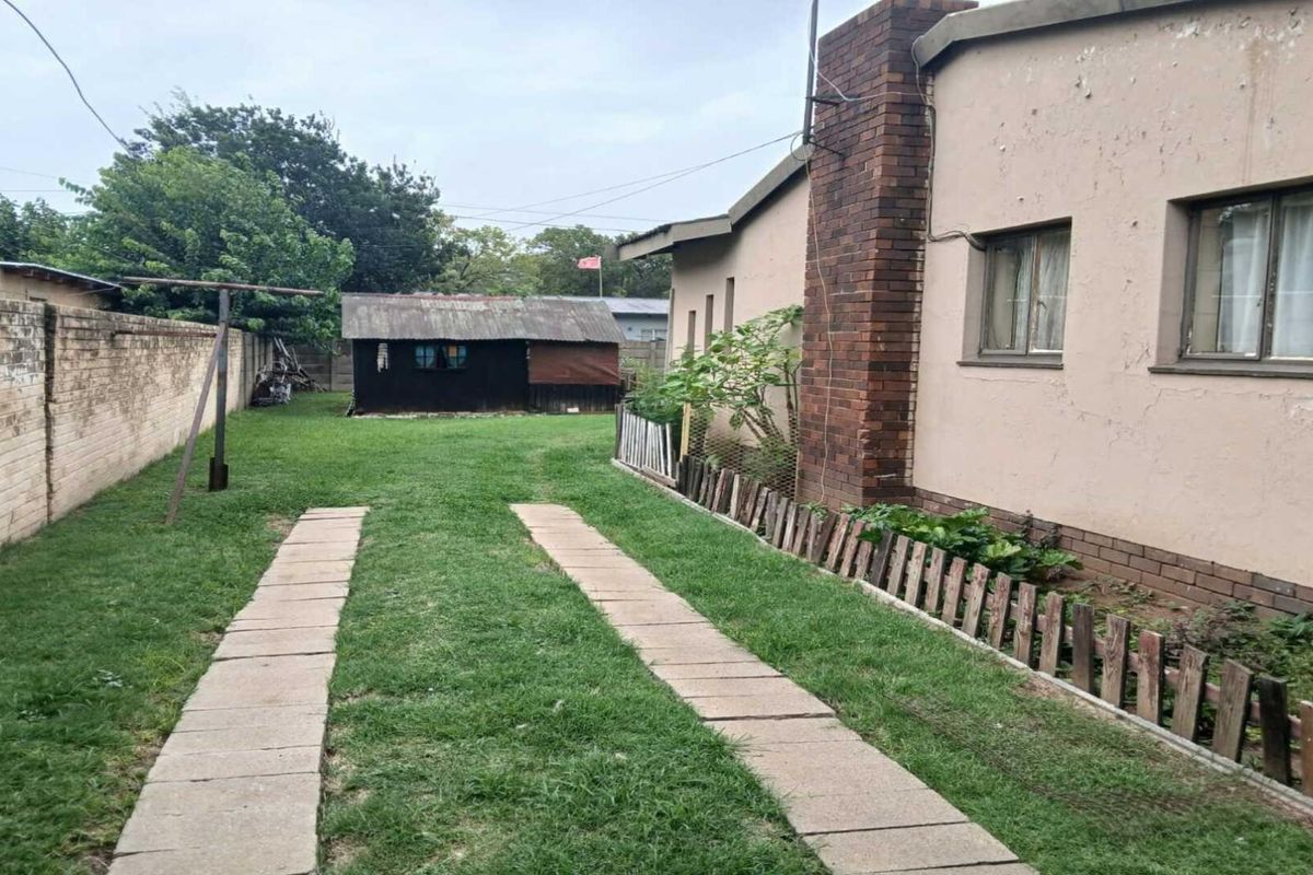 A view of the rear garden featuring a lawn, paved driveway tracks, a brick chimney, and a detached outbuilding structure.