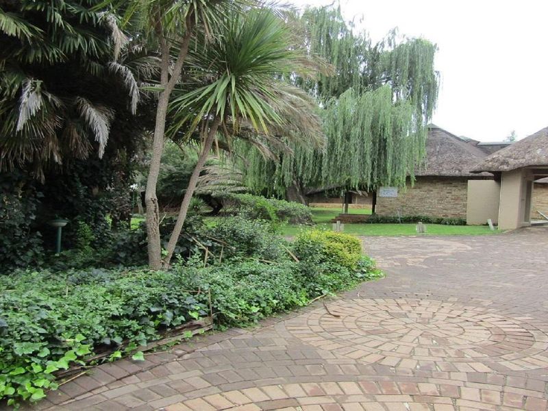 A view of the property entrance featuring a paved driveway, established garden with mature trees, and a thatched roof building structure.